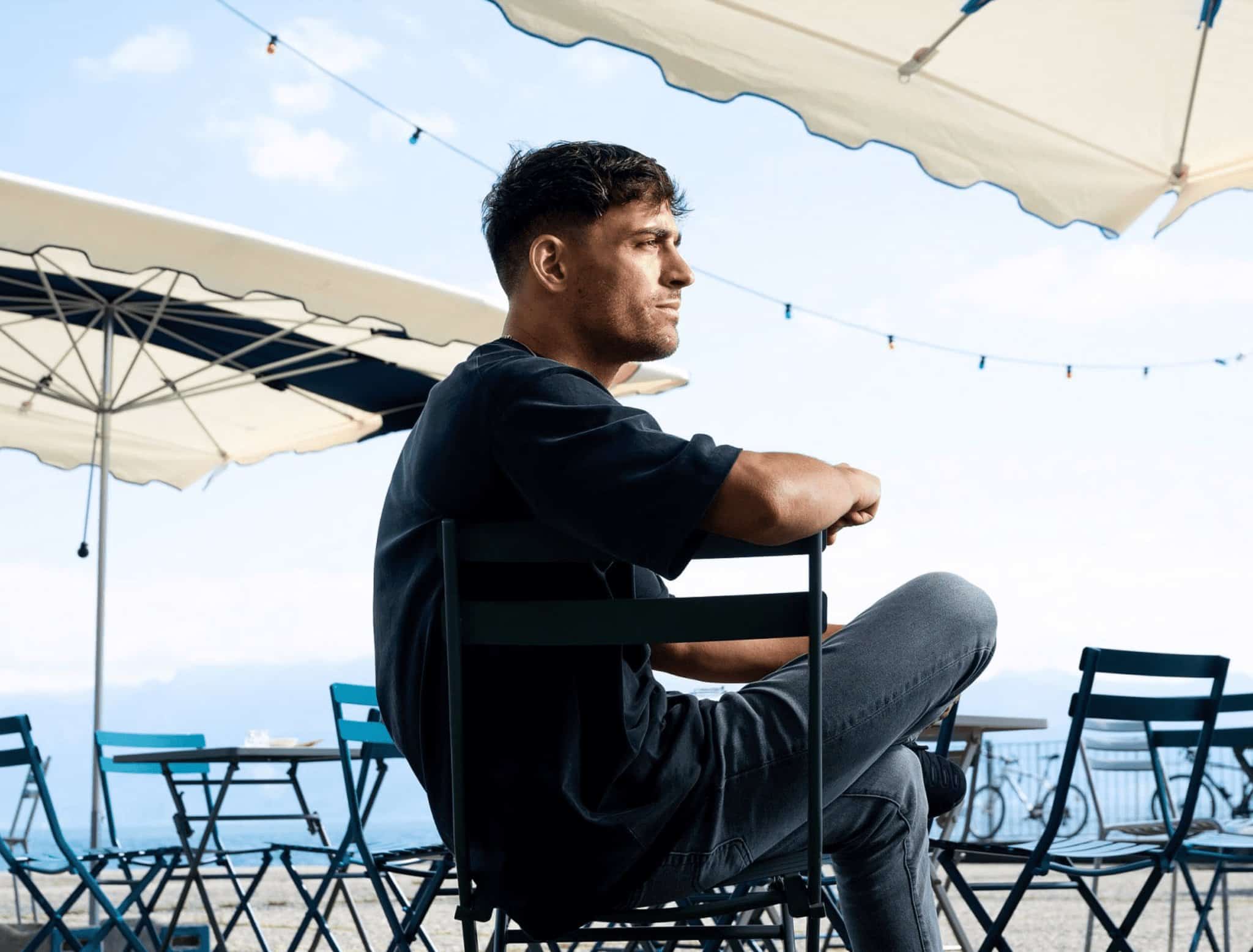 Jeune homme pensif assis en terrasse avec parasols et vue sur la mer.